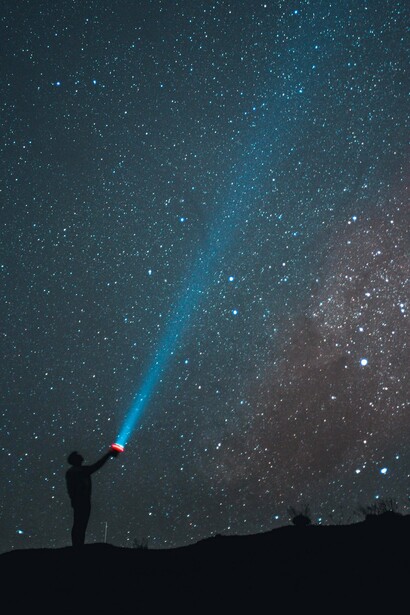 Uma silhueta aponta uma lanterna para o céu coberto de estrelas durante a noite. Tal como os escritores citados no texto, o gesto parece procurar respostas na vastidão infinita