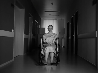 In the hospital corridor, a patient sits in a wheelchair, their presence marked by a quiet resilience as they observe the bustling surroundings