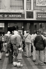 A black-and-white photograph of people standing in a queue outside a shop, Belgium