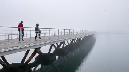 Pedestrians crossing a long wooden bridge over calm water on a misty, fog-covered morning