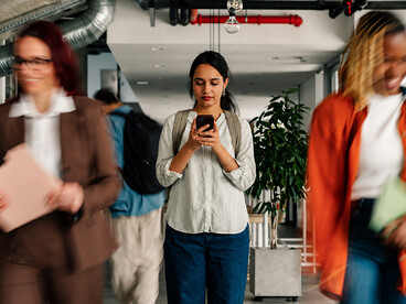 A young woman focuses on her smartphone while moving through a bustling office corridor, seemingly unaware of the people passing by