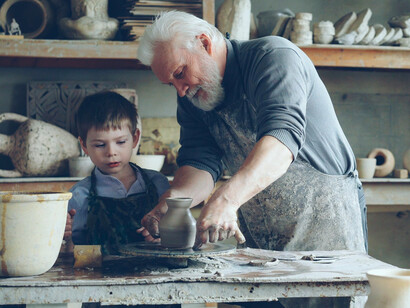 An elderly man mentoring a child as he creates pottery on a spinning wheel, reflecting the transfer of skills, wisdom, and tradition across generations