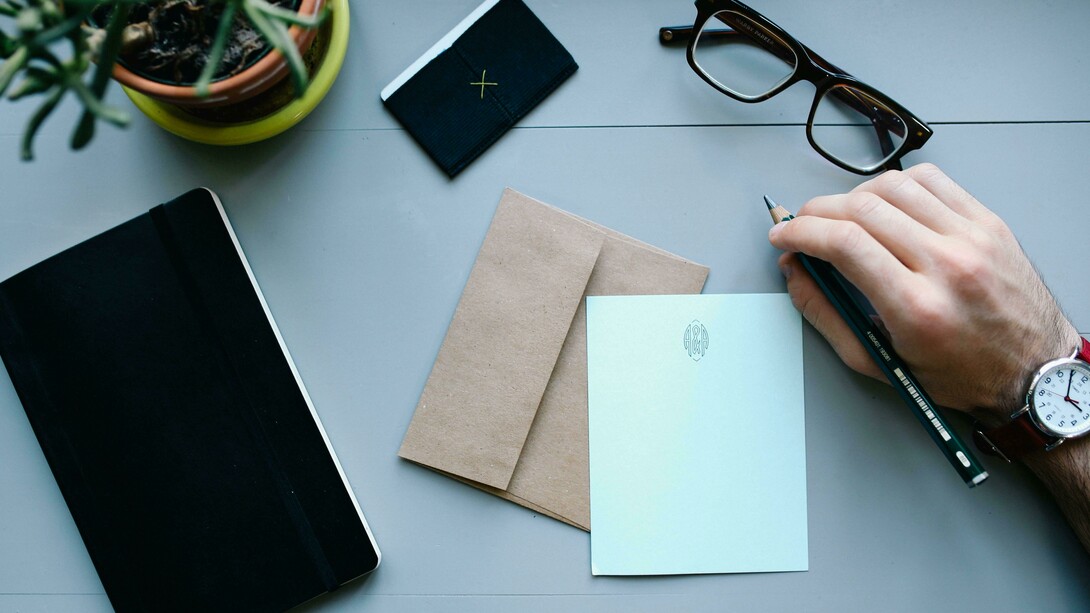 A person writing at a desk with a notebook and plant on