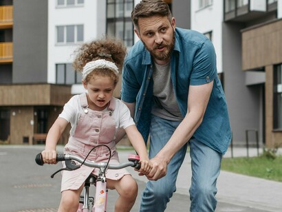 A father helps his daughter learn to ride a bike, their laughter and smiles reflecting the pure joy of shared moments, illustrating how such experiences enrich our lives far beyond the demands of overworking and constant busyness