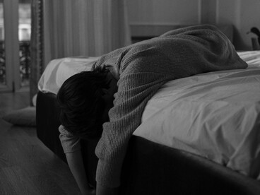 A solitary figure sits under a blanket, resting her head in her hand, reflecting a state of anxiety