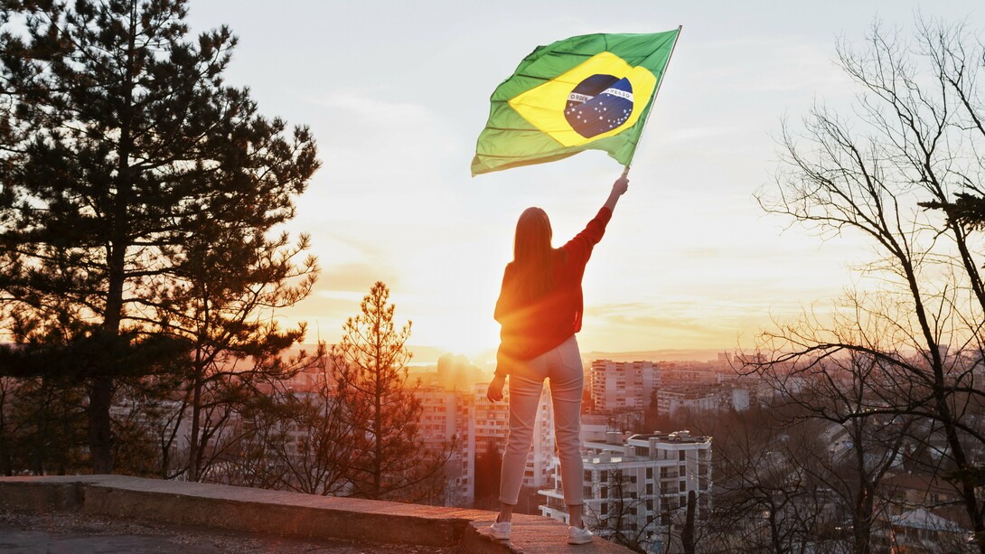 Uma mulher de calça branca e camisa vermelha segura a bandeira brasileira diante de uma cidade iluminada pelo sol. Mais do que um símbolo nacional, a cena sugere a presença feminina como parte ativa das escolhas que definem os caminhos do Brasil