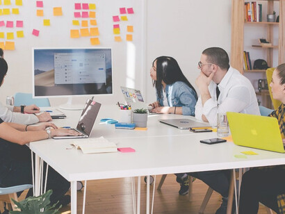 Seated around a table, five individuals gaze intently at a turned-on white iMac, engrossed in its vibrant display, suggesting a productive and collaborative work environment