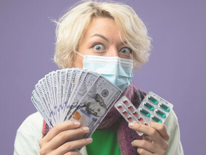 A woman with short hair, wearing a warm scarf and a facial protective mask, looks shocked as she holds cash and a blister pack of pills, illustrating the economic challenges of healthcare costs