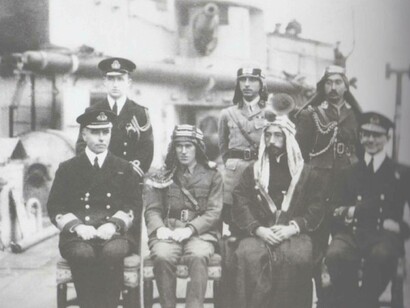 British officer Thomas Edward Lawrence (center) is sitting in his officer's uniform but wearing an Arabian head scarf and is seated next to Emir Faisal I 
