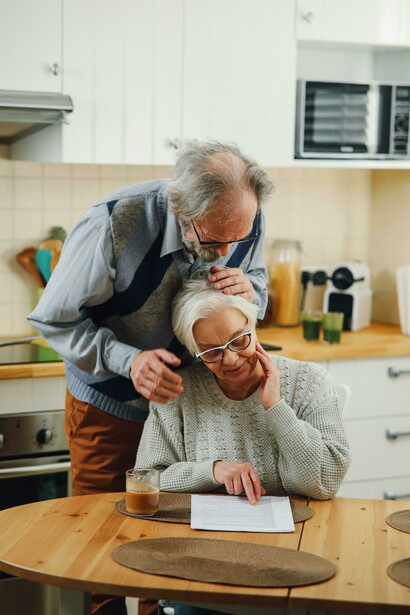 An elderly couple sits in the kitchen, reading a document and laughing together, illustrating the comfort and connection of a long-term relationship