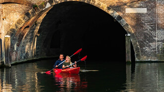 A man and woman paddle a red kayak along a river in Utrecht, the Netherlands, under the bright daytime sky