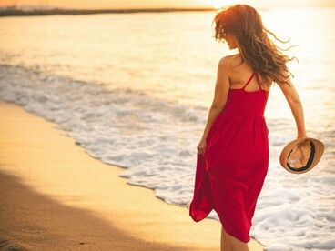 De costas, uma mulher de vestido vermelho e chapéu de palha caminha pela praia, junto ao mar. Afastar-se por alguns instantes do ruído cotidiano pode abrir espaço para escutar a si mesmo e reconhecer o caminho que realmente faz sentido seguir