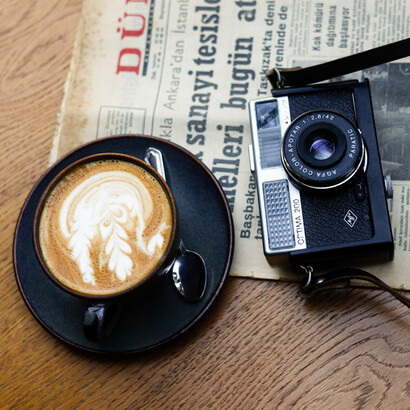 Un periódico, una cámara y una taza de café se encuentran sobre una mesa, como una pequeña escena del oficio periodístico: informarse, mirar y contar