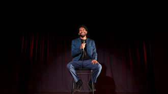 A man performing stand-up comedy on stage in a lively comedy club atmosphere