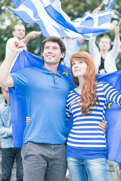 A Greek couple stands side by side during a protest, holding Greek flags as a symbol of resistance against home foreclosures and financial injustice
