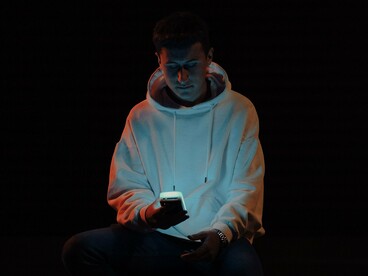 Um jovem de moletom aparece sentado na escuridão, concentrado no smartphone que ilumina seu rosto. A cena transforma um gesto cotidiano em uma imagem de introspecção, na qual a tela parece ocupar quase todo o campo de atenção. Estar constantemente conectado não significa necessariamente estar envolvido com o que vivemos: reconhecer o tédio pode ser o primeiro passo para compreender o que buscamos ao recorrer repetidamente ao celular.