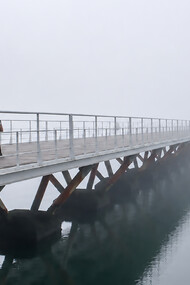 Pedestrians crossing a long wooden bridge over calm water on a misty, fog-covered morning