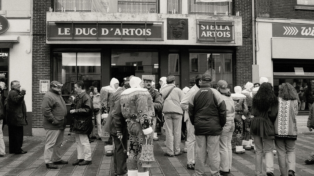 A black-and-white photograph of people standing in a queue outside a shop, Belgium