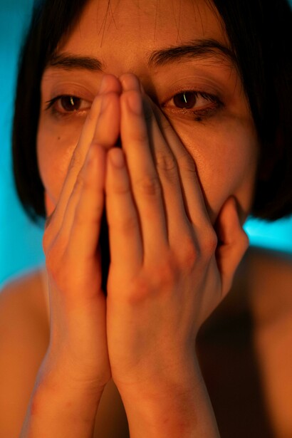 A short-haired woman in a white tank top rests her hand on her face, looking upset