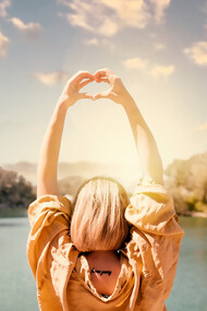 Experience a symbolic moment of self-discovery as a woman creates heart-shaped hands against a sunset backdrop in this captivating image