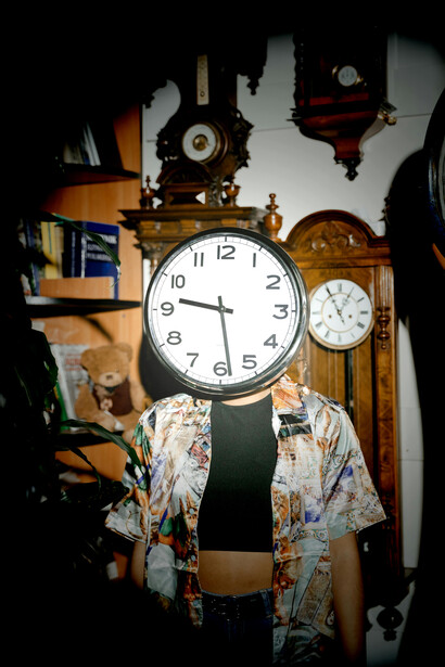 A person stands in front of wooden shelves filled with clocks, holding one over their face—symbolizing the loss of identity in a time-pressured, accelerated society