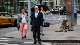 Busy city intersection in the USA highlighting social inequality, as pedestrians move through an urban street while a homeless woman sits unnoticed on the sidewalk