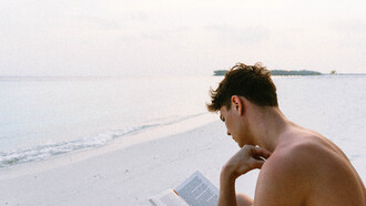 Rapaz lê livro em beira da praia de frente para o mar