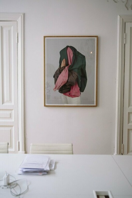 Parede branca com um quadro. Mesa e cadeira brancas com alguns papeis