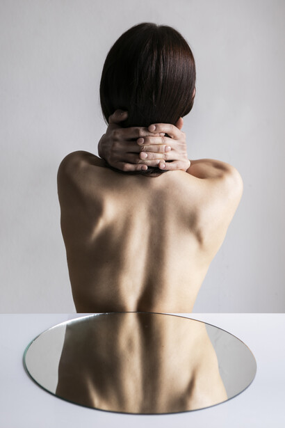 Ritratto di schiena di una donna, con le mani che le circondano il collo, il cui dorso è riflesso nello specchio