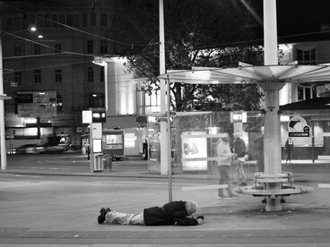 Um homem está deitado no chão em uma cena noturna registrada em preto e branco, nas proximidades de uma parada de ônibus. A imagem conduz à reflexão sobre as pessoas cuja vulnerabilidade permanece presente nos espaços públicos, mesmo quando passa despercebida