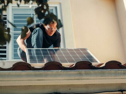 A man installs a solar panel while standing on the roof of a house