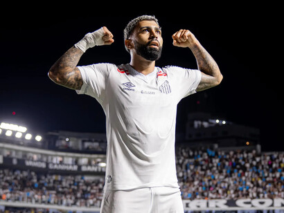 Com os dois braços flexionados e os punhos erguidos, Gabigol olha para cima vestindo a camisa branca do Santos, enquanto a torcida aparece ao fundo, nas arquibancadas iluminadas. O gesto transmite a intensidade emocional de quem vive o futebol dentro de campo. De volta ao clube que o revelou, o atacante tem a oportunidade de transformar a expectativa pelo seu retorno em gols, boas atuações e uma nova aproximação com os torcedores santistas