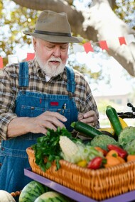 An elderly man wearing denim overalls and a hat, examining fresh vegetables