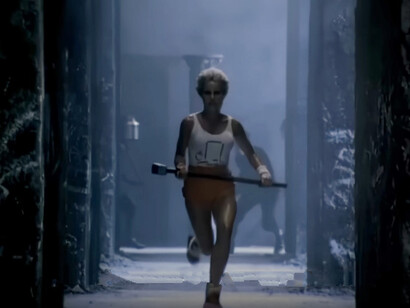 Nella pubblicità, una giovane donna atletica, vestita di rosso e bianco, corre verso uno schermo gigante e lo distrugge con un martello, simboleggiando la rottura con il pensiero unico e l'innovazione portata da Apple