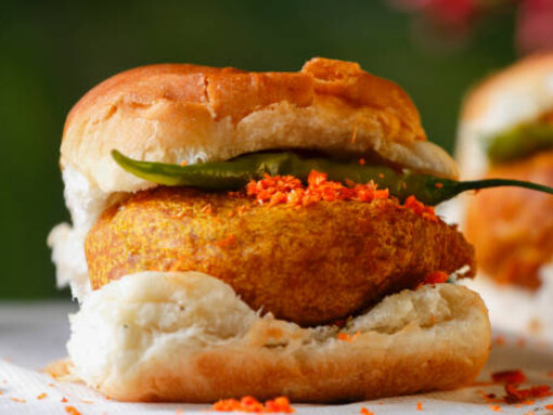 Restaurant scene featuring a plate of Mumbai Vada Pav—deep-fried potato dumpling in a bread bun topped with green chili and tomato. In the foreground, an unrecognizable person picks up a slice of tomato. The focus is on the plate, set in an Indian setting