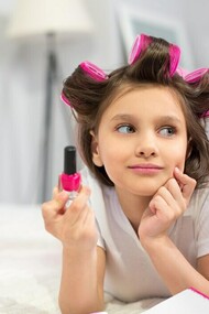 Preschool-aged girl lying on a white blanket with hair curler, holding nail polish 