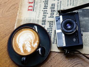 Un periódico, una cámara y una taza de café se encuentran sobre una mesa, como una pequeña escena del oficio periodístico: informarse, mirar y contar