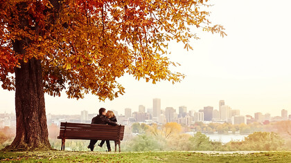 A young married couple sits on a bench, sharing a kiss as the man lovingly wraps his arm around his wife’s shoulders on a beautiful autumn day