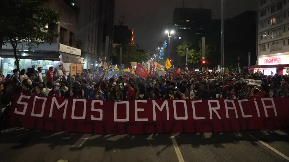 Uma multidão ergue a faixa “somos democracia” durante protesto popular. A disputa entre participação democrática e projetos de poder autoritários está no centro das tensões políticas contemporâneas