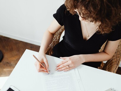 As mãos de uma mulher assinam um documento, transformando uma ação burocrática em um gesto de responsabilidade. No artigo, enquanto a mãe permanece como referência concreta, o lugar reservado ao pai continua sendo apenas um campo em branco — uma ausência que o papel registra, mas não explica