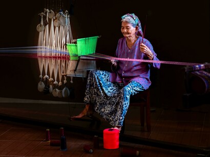 Uma artesã trabalha manualmente num tear tradicional rodeada por fios coloridos e instrumentos de tecelagem. Antes da industrialização, a produção de tecidos dependia de técnicas artesanais transmitidas entre gerações
