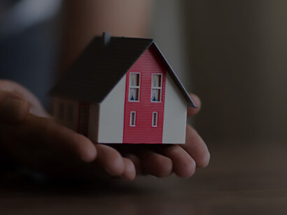 Amidst America's housing crisis and economic downturn, a businessman's hand grips a miniature house savings model, symbolizing the struggle and hope in navigating the current real estate climate