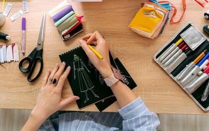 Mãos desenhando roupa em um caderno sobre uma mesa de estudos de moda. Entre traços e ideias, nasce a moda em um processo criativo que une arte, técnica e consciência em cada detalhe. O primeiro esboço de uma peça carrega mais que estética, revela pensamento, cultura e intenção. Antes das passarelas, a moda começa no papel, onde criatividade e propósito se encontram