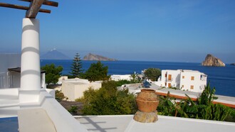 Le architetture vernacolari insulari delle Isole Eolie si caratterizzano per l’uso di materiali locali come pietra lavica e calce, adattandosi perfettamente al clima e al paesaggio mediterraneo. La foto (di Dylan Parker) mostra una tipica costruzione di Panarea sul panorama del Mar Mediterraneo 