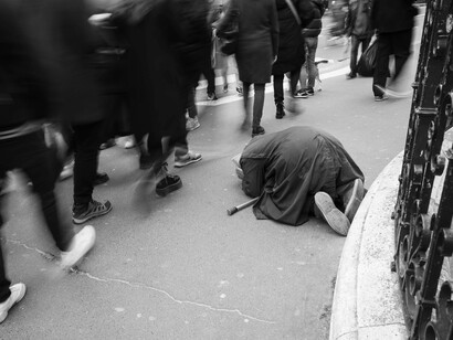 Em uma cena urbana em preto e branco, uma pessoa permanece no chão enquanto a multidão se movimenta ao redor. O contraste entre a imobilidade de quem está vulnerável e a pressa cotidiana dos demais convida à reflexão sobre a indiferença diante do sofrimento alheio
