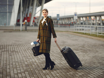 A stylish woman walks near a contemporary building with her luggage, passport, and boarding ticket in hand