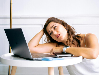 A woman wearing a smartwatch and holding a smartphone sits beside a laptop, appearing overwhelmed by media overload