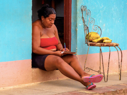 A local woman in South America sits on the floor, writing thoughtfully in her notebook, capturing the stories and traditions of her vibrant community