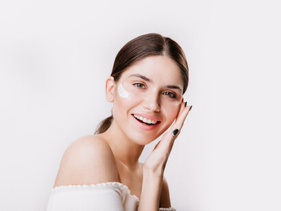 A smiling girl with green eyes applies cream to her clean face, embracing self-care and using beauty products designed for acne treatment