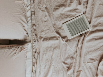 Um leitor de livros digitais ocupa o centro de uma cama coberta por tecidos claros, em uma composição marcada pela simplicidade e pelo conforto. A leitura contemporânea encontra no ambiente digital uma nova forma de preservar a experiência de mergulhar em uma história